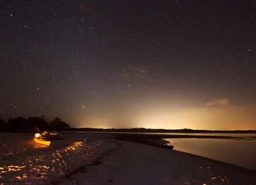 A serene night scene by a river, featuring a starry sky and a glowing campfire on the sandy shore.