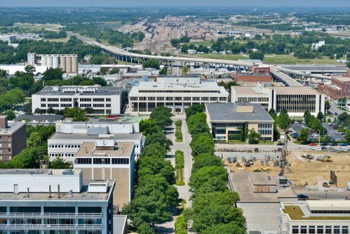 Aerial view of a cityscape featuring buildings, trees, and a railway in the background under a clear blue sky.