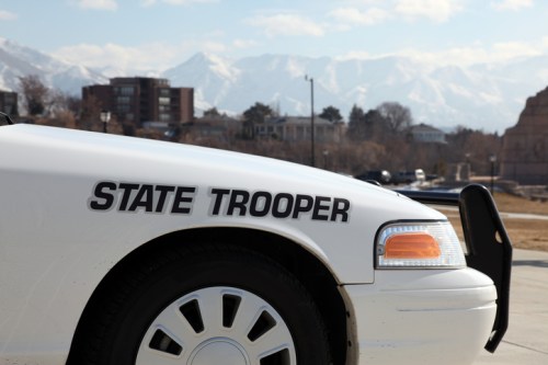 Close-up of a white police car with "STATE TROOPER" written on the side, mountains in the background.