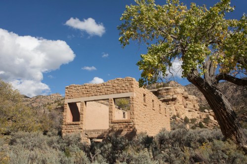 Ruins of a stone building surrounded by shrubs and trees, set against a blue sky with fluffy clouds.