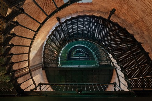 A spiral staircase viewed from above, featuring metal steps and a brick wall background.