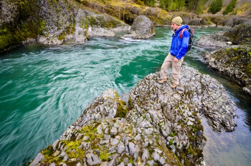 A person in a blue jacket stands on a rocky outcrop by a flowing river, surrounded by lush greenery.