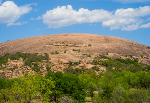 A large, smooth rock formation rises against a blue sky with fluffy clouds, surrounded by green vegetation.