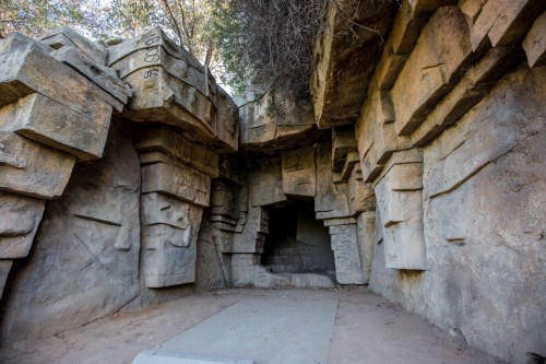 A rocky cave entrance with angular stone formations and overhanging vegetation.