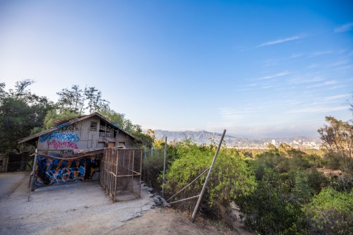 A weathered house with graffiti, surrounded by greenery, overlooking a distant city skyline and mountains.