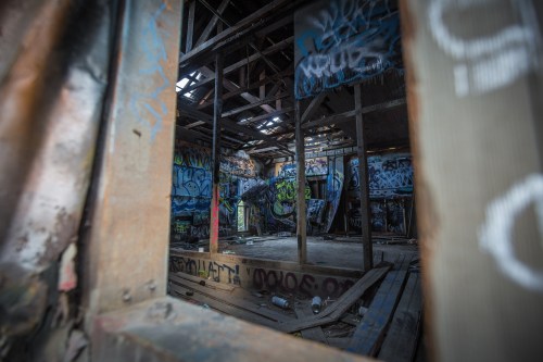 View through a broken window into a graffiti-covered, abandoned building with exposed wooden beams and debris on the floor.