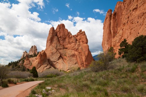 Rocky formations rise against a blue sky with clouds, surrounded by green grass and trees along a winding path.