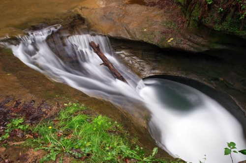 A serene waterfall cascades over rocks, with a smooth flow and a log resting in the water, surrounded by lush greenery.