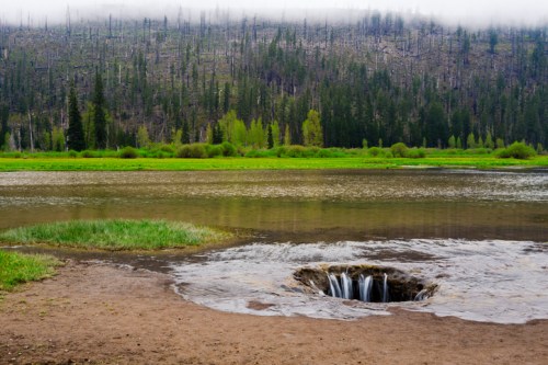 A serene landscape featuring a small waterfall flowing into a circular hole, surrounded by lush greenery and a forested hillside.