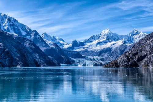 A serene landscape featuring snow-capped mountains reflecting in calm blue waters, with a glacier in the background.