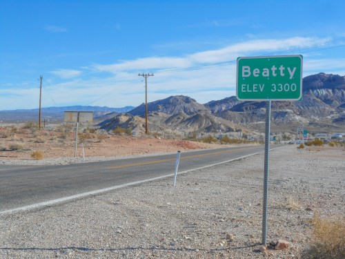 A green sign for Beatty, elevation 3300, beside a road with mountains in the background under a clear blue sky.