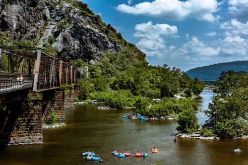 A scenic river view with people floating on tubes, surrounded by lush greenery and rocky cliffs under a blue sky.