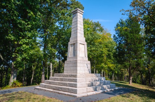 A tall stone monument surrounded by trees on a sunny day, featuring a stepped base and a rectangular top.