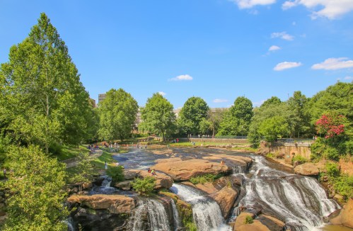 A scenic view of a waterfall surrounded by lush greenery and people enjoying the outdoors under a clear blue sky.