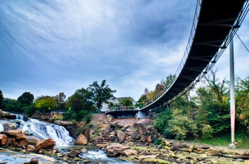 A suspension bridge arches over a waterfall, surrounded by lush greenery and a cloudy sky.
