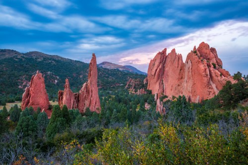 A scenic view of red rock formations surrounded by greenery under a blue sky with clouds at dusk.