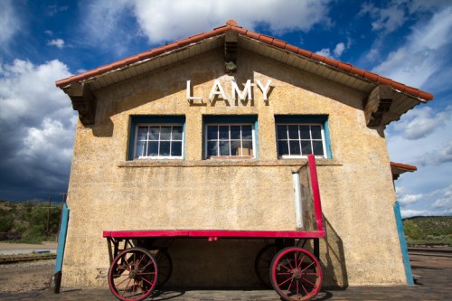 Historic Lamy train station with a red cart in front, set against a cloudy blue sky.