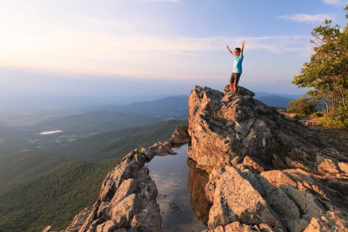 A person stands triumphantly on a rocky outcrop, overlooking a lush valley and mountains under a clear sky.