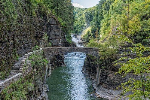 A stone bridge spans a river surrounded by steep, rocky cliffs and lush green trees under a bright blue sky.