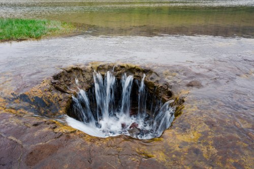 A circular rock formation with cascading water flowing into a deep hole, surrounded by grass and a calm river.