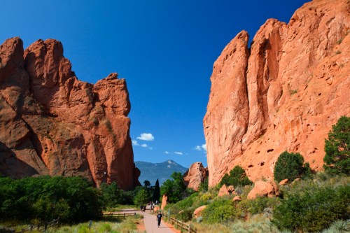 A scenic view of red rock formations with a clear blue sky, featuring a walking path and lush greenery.