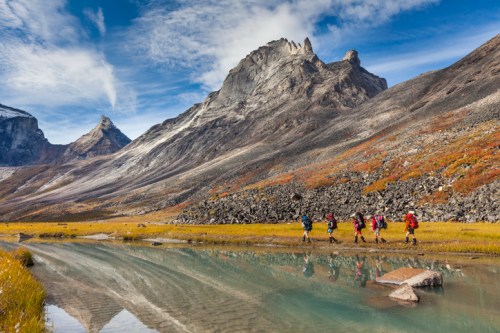 A group of hikers walks along a river in a mountainous landscape with colorful autumn foliage and clear blue skies.