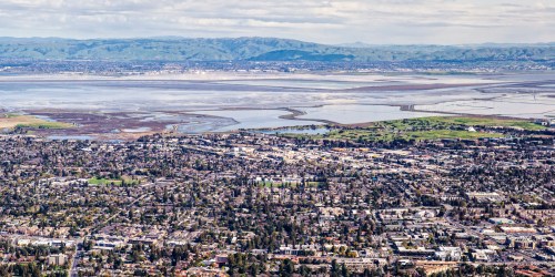 Aerial view of a city with green hills and a bay in the background, showcasing urban and natural landscapes.