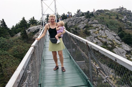 A woman in a black tank top and green skirt carries a child while walking on a suspension bridge surrounded by trees.