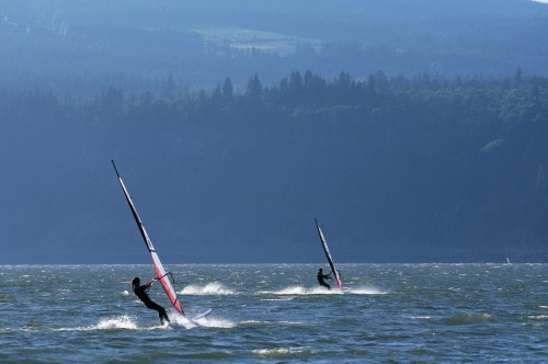 Two windsurfers glide across a lake, surrounded by misty mountains and lush green trees in the background.