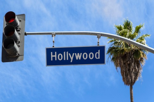 A street sign reading "Hollywood" hangs above a traffic light, with a palm tree in the background against a blue sky.