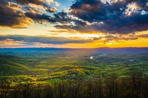 A scenic view of rolling green hills under a colorful sunset with clouds and a winding river in the distance.