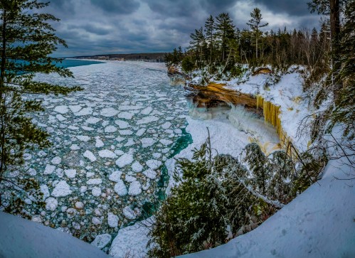 A winter landscape featuring a frozen lake with ice floes, snow-covered cliffs, and evergreen trees under a cloudy sky.