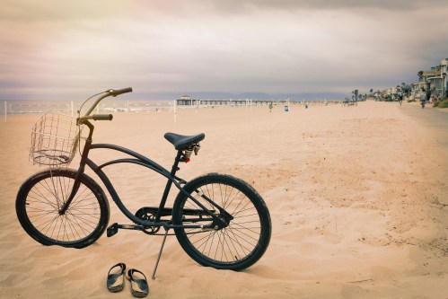 A black bicycle with a basket rests on sandy beach, with a cloudy sky and distant pier in the background.