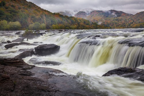 A scenic view of a waterfall cascading over rocky terrain, surrounded by lush, colorful autumn foliage and misty mountains.