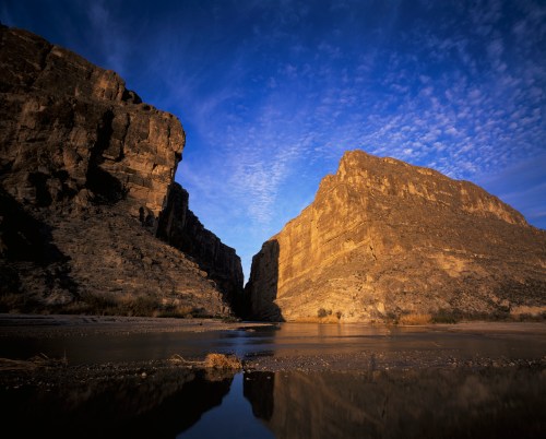 A serene landscape featuring towering cliffs reflecting in a calm river under a blue sky with scattered clouds.