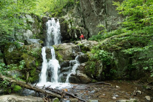 A serene waterfall cascades over rocks, surrounded by lush greenery, with a person in red sitting nearby.