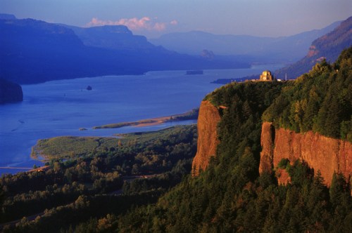 A scenic view of a river winding through mountains, with a cliffside structure and lush greenery in the foreground.
