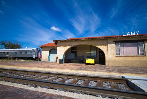 A train station with a yellow cart, an old train, and a blue sky with clouds in Lamy, New Mexico.
