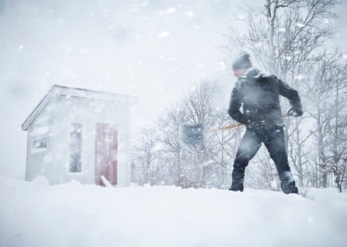 A person shoveling snow in a heavy snowstorm near a small shed surrounded by trees. Visibility is low due to the snow.