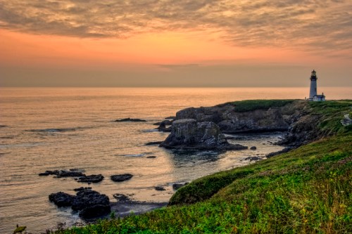 A lighthouse stands on a rocky coastline at sunset, with calm waters and a colorful sky in the background.