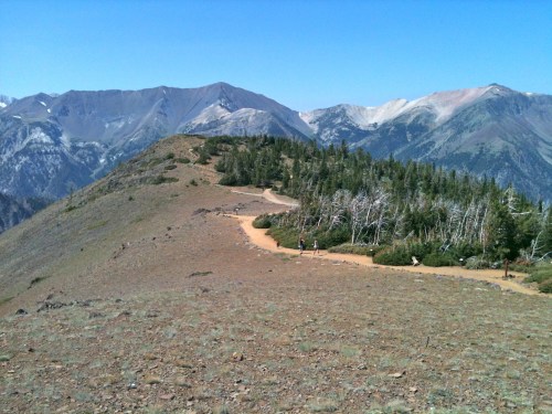 A winding trail leads through a mountainous landscape with sparse trees and rocky terrain under a clear blue sky.