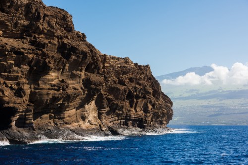 Rocky coastline with steep cliffs, clear blue water, and distant mountains under a bright sky with scattered clouds.