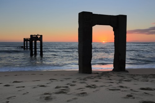 Silhouetted pier remnants at sunset, with waves gently lapping the sandy beach and a colorful sky in the background.
