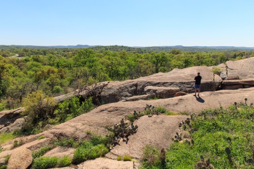 A person stands on a rocky outcrop overlooking a lush green landscape under a clear blue sky.