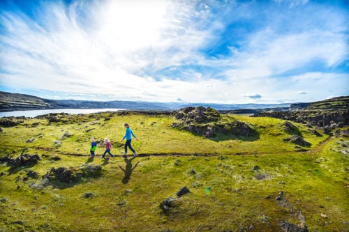 A person and two children walk along a grassy path with a scenic landscape and blue sky in the background.