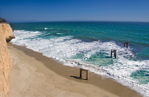 A sandy beach with cliffs, gentle waves, and remnants of wooden structures partially submerged in the water.