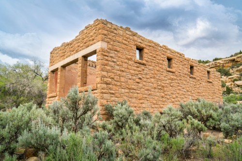 A weathered stone building surrounded by shrubs, set against a cloudy sky in a natural landscape.