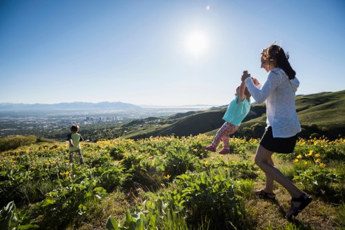 A woman swings a child in a sunny hillside field, with a city skyline and mountains in the background.