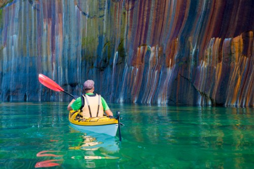 A kayaker paddles in clear water near colorful, striped rock formations.