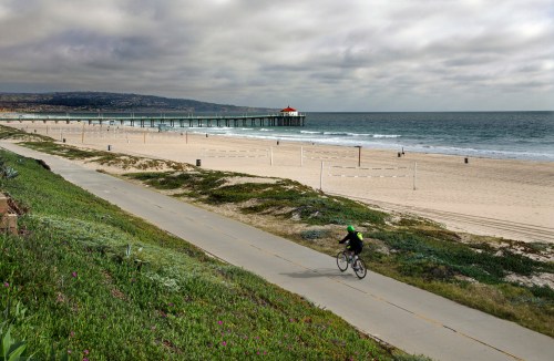 A cyclist rides along a beach path with a pier in the background under a cloudy sky.